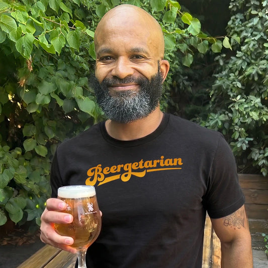 A smiling bald man with a gray beard holds a glass of beer outdoors while wearing a black t-shirt with the word 'Beergetarian' printed in orange across the chest.