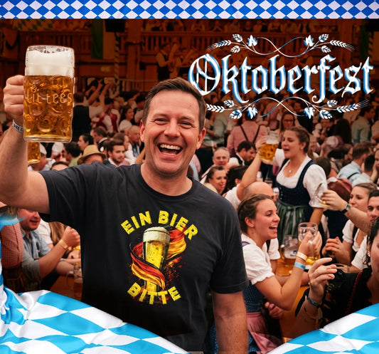 A smiling man in a beer hall raises a large mug of beer while wearing a black graphic T-shirt that reads 'EIN BIER BITTE' with a beer illustration. The background shows a crowded indoor Oktoberfest scene with people, beer mugs, and a blue-and-white checkered banner and sign at the top right.