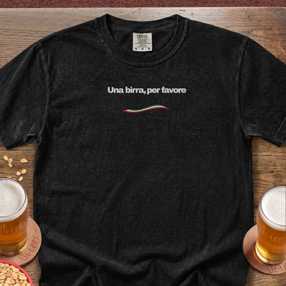 A black short-sleeve t-shirt with the Italian phrase 'Una birra, per favore' printed in white across the chest, above a small red, white, and green wave. The shirt is laid flat on a wooden table with beer glasses and peanuts as props.
