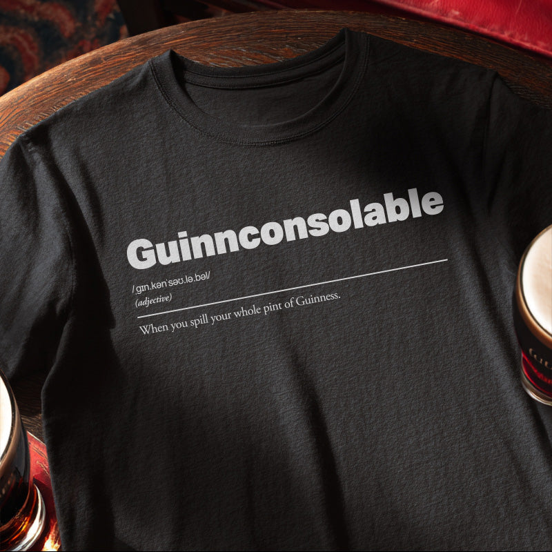 Black t-shirt with 'Guinnconsolable' text design, placed on a wooden surface with a pint of Guinness. Guinnconsolable :: When you spill your whole pint of Guinness.