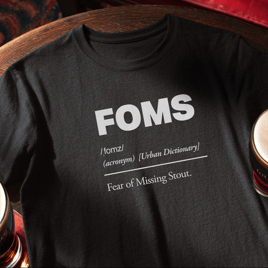 Black t-shirt with 'FOMS - Fear of Missing Stout' definition text on a wooden pun table with pints.