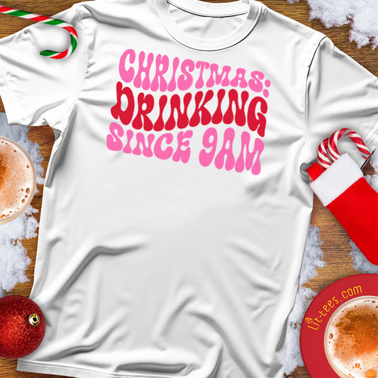White t-shirt with pink text 'Christmas Drinking Since 9am' on a festive background with coffee cups and stockings.