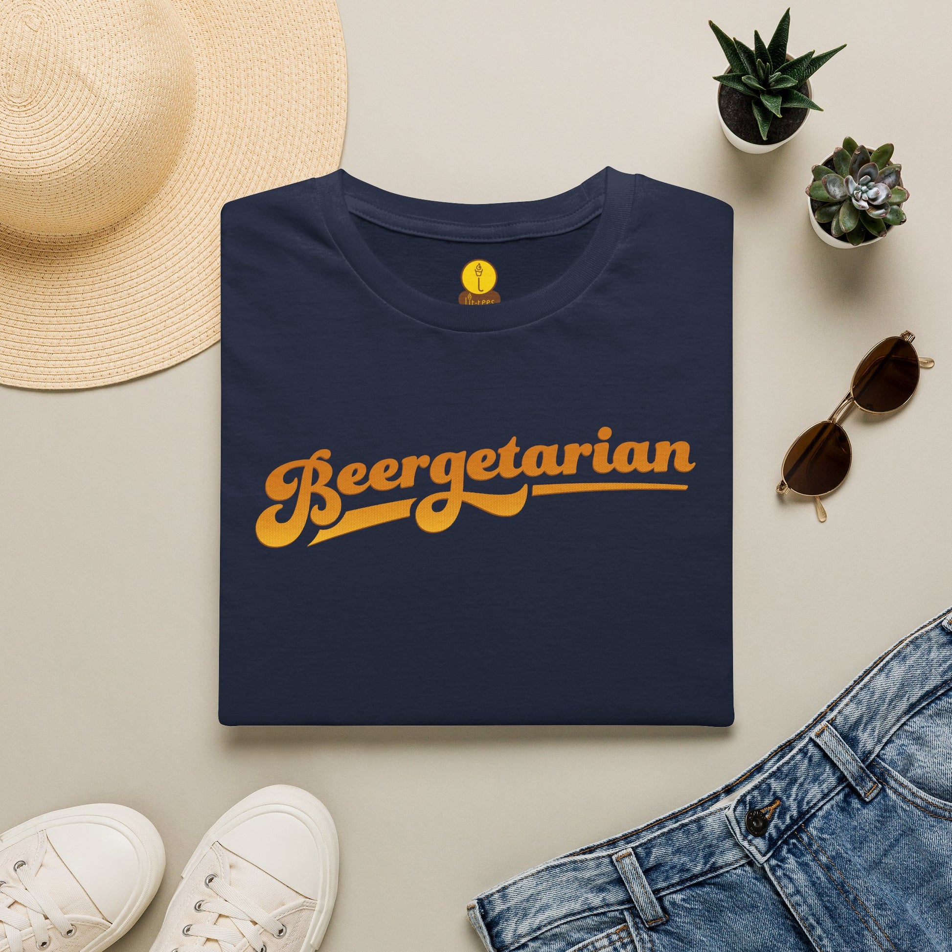A folded navy blue t-shirt with 'Beergetarian' printed in a retro orange-yellow font across the chest, styled in a flat lay with a straw hat, white sneakers, sunglasses, a pair of blue jeans, and small potted succulents on a light beige background.