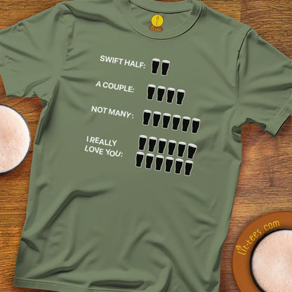 An Military Green green crewneck t-shirt with a humorous beer-themed graphic, laid flat on a wooden surface next to two pints of beer. The graphic reads 'SWIFT HALF: 2 glasses, A COUPLE: 4 glasses, NOT MANY: 8 glasses, I REALLY LOVE YOU: 16 glasses'.