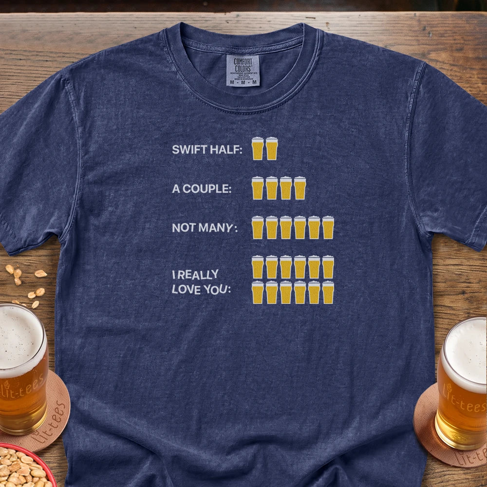 A navy blue graphic t-shirt laid flat on a wooden table, featuring a humorous beer-themed print with text and beer glass illustrations. The shirt is styled with glasses of beer and peanuts on a rustic wooden background.