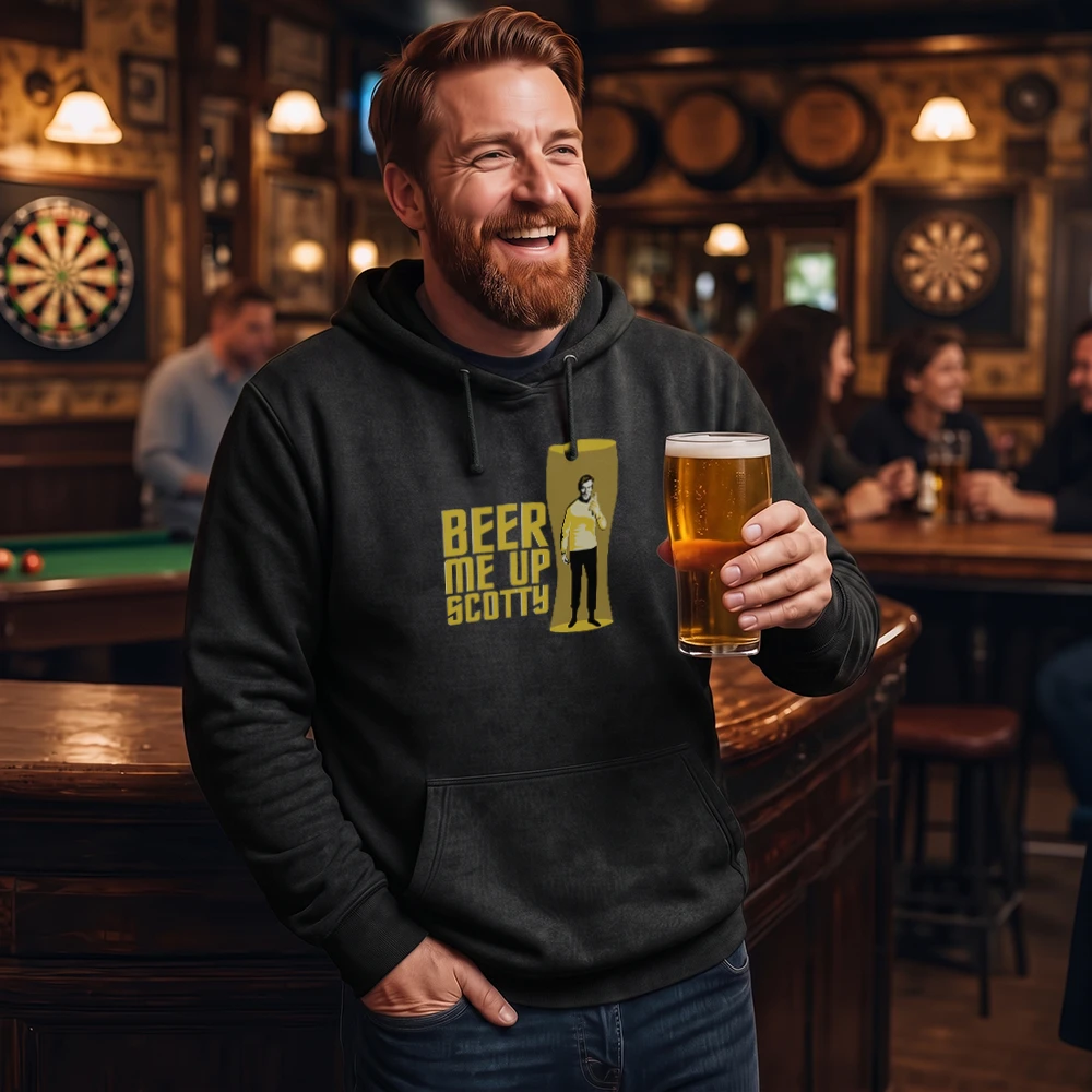 A smiling man in a bar wearing a black hoodie with a yellow graphic that reads 'BEER ME UP SCOTTY' and holding a glass of beer. The background shows a dimly lit pub interior with other patrons, dartboards, and warm lighting.