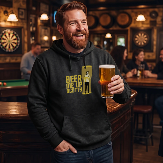A smiling man in a bar wearing a black hoodie with a yellow graphic that reads 'BEER ME UP SCOTTY' and holding a glass of beer. The background shows a dimly lit pub interior with other patrons, dartboards, and warm lighting.