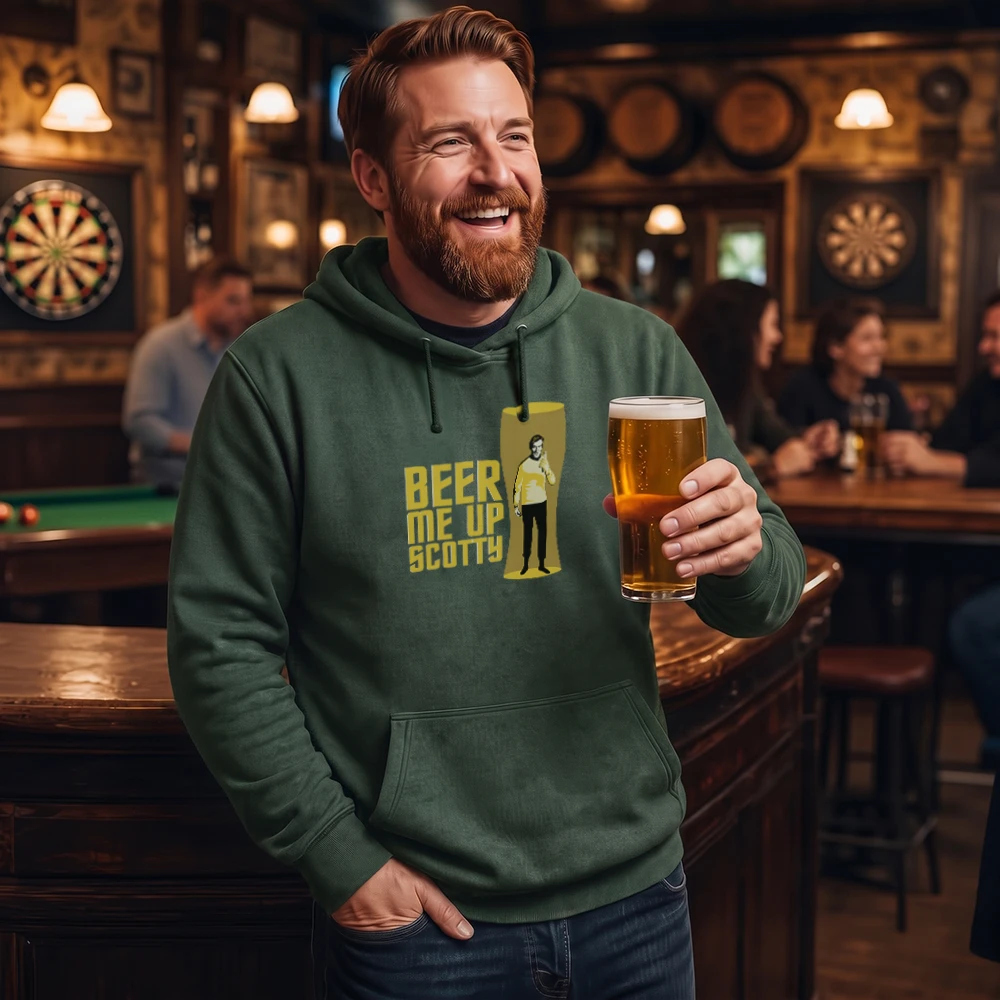 A smiling man in a bar wearing a dark green hoodie with a yellow graphic that reads 'BEER ME UP SCOTTY' and holding a pint of beer. The background shows a dimly lit pub interior with dartboards, lamps, and other patrons.