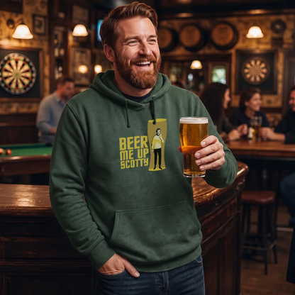 A smiling man in a bar wearing a dark green hoodie with a yellow graphic that reads 'BEER ME UP SCOTTY' and holding a pint of beer. The background shows a dimly lit pub interior with dartboards, lamps, and other patrons.