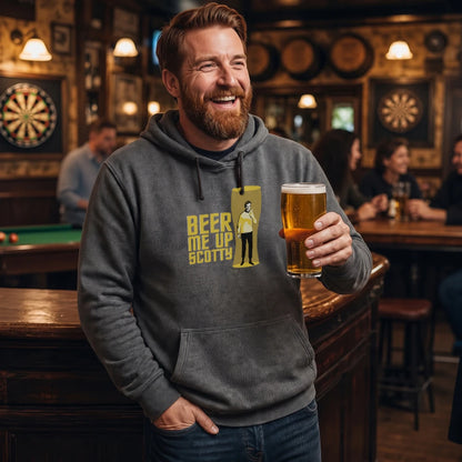 A smiling man in a bar wearing a dark gray hoodie with a yellow graphic that reads 'BEER ME UP SCOTTY' next to a figure silhouette. He is holding a pint of beer in his hand, with a blurred pub interior in the background.