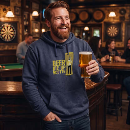 A smiling man in a bar wearing a navy hoodie with a yellow graphic that reads 'BEER ME UP SCOTTY' and holding a pint of beer. The background shows a dimly lit pub interior with other patrons, tables, and dartboards.