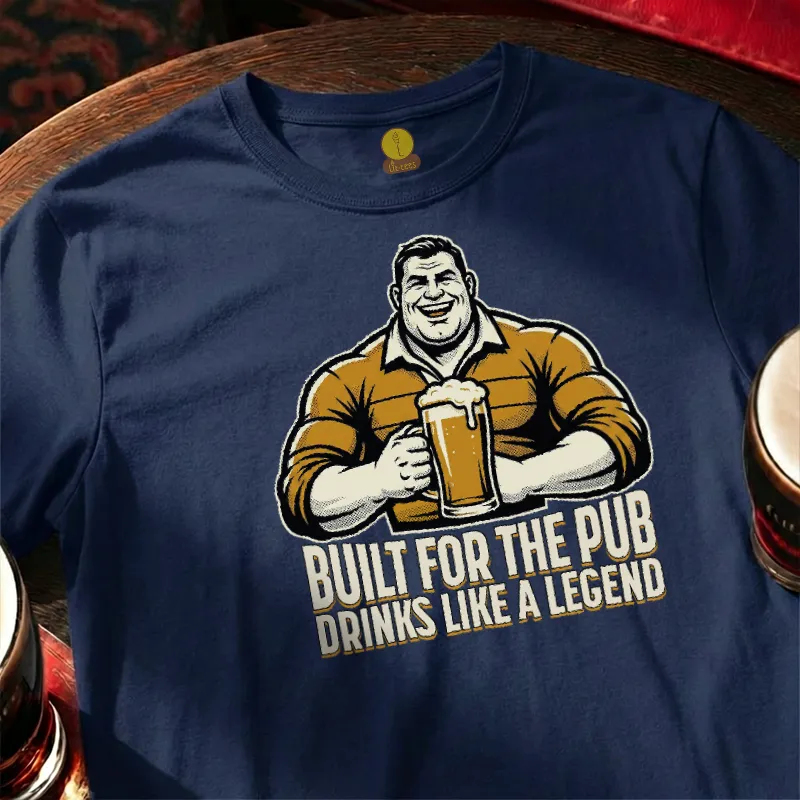 Navy blue t-shirt with graphic of a man holding a beer mug and text 'Built for the pub drinks like a legend' on a wooden bar table