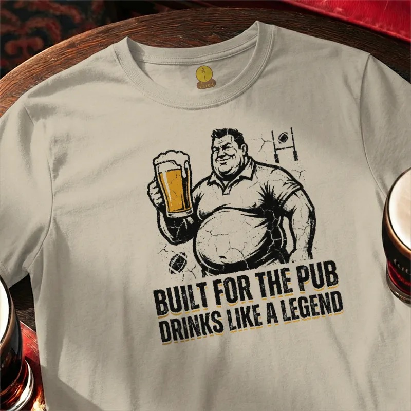T-shirt with a graphic of a muscular character holding a beer and text 'Built for the pub drinks like a legend' on a wooden bar table