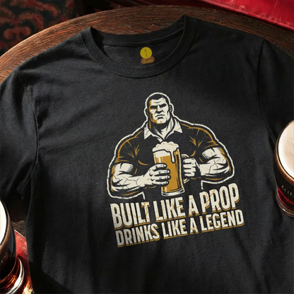 Black t-shirt with graphic of a man holding a beer mug and text 'Built Like a Prop Drinks Like a Legend' on a wooden bar table
