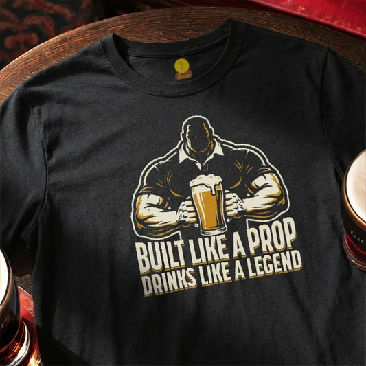 Black t-shirt with graphic design and text 'Built Like a Prop Drinks Like a Legend' on a wooden bar table.