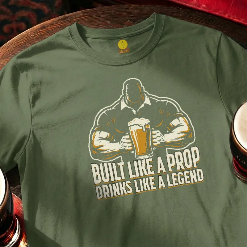 Green t-shirt with graphic of a man holding a beer and text 'Built Like a Prop Drinks Like a Legend' on a wooden bar table.