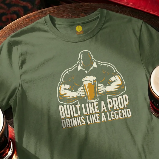 Green t-shirt with graphic of a man holding a beer and text 'Built Like a Prop Drinks Like a Legend' on a wooden bar table.
