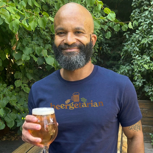A smiling bald man with a gray beard holds a glass of beer outdoors, wearing a navy blue t-shirt with the word 'beergetarian' printed in orange-brown across the chest.