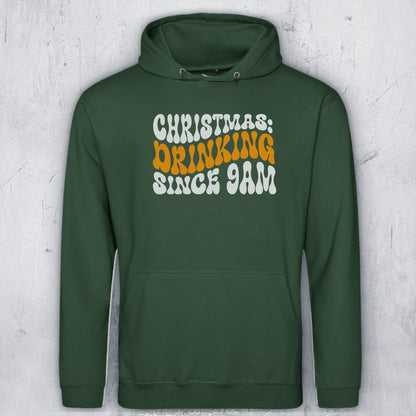 A dark green pullover hoodie with a front graphic reading 'CHRISTMAS DRINKING SINCE 9AM' in white and orange lettering, displayed against a light gray textured background.