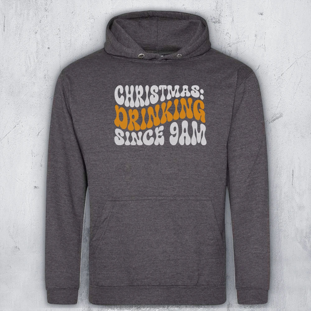 A dark heather gray pullover hoodie with a front graphic that reads 'CHRISTMAS DRINKING SINCE 9AM' in white and orange lettering, displayed against a light gray textured wall.