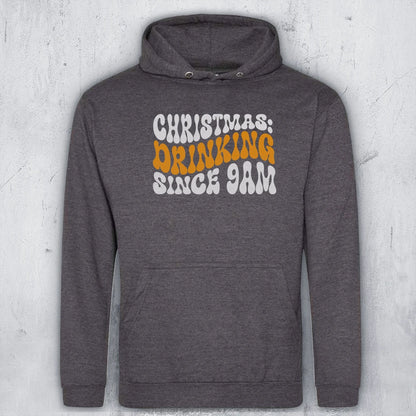 A dark heather gray pullover hoodie with a front graphic that reads 'CHRISTMAS DRINKING SINCE 9AM' in white and orange lettering, displayed against a light gray textured wall.