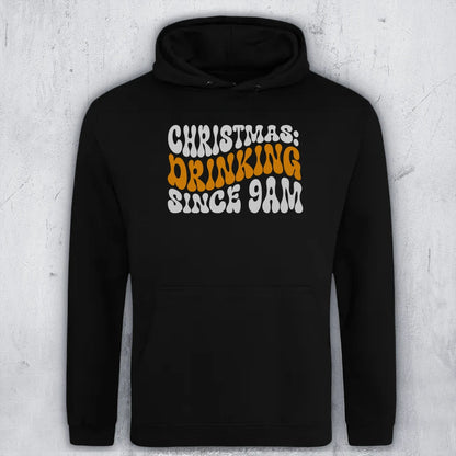 A black pullover hoodie with a front graphic reading 'CHRISTMAS: DRINKING SINCE 9AM' in white and orange retro-style lettering, shown against a light gray textured wall.