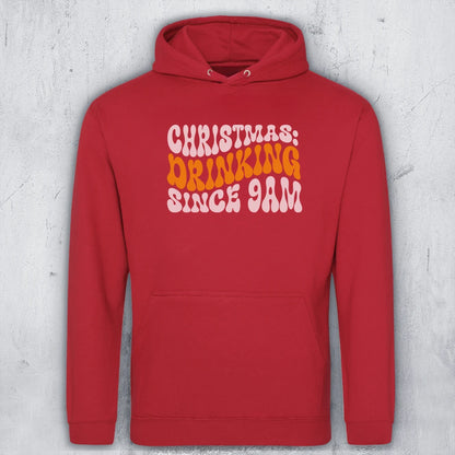 A red pullover hoodie with a front kangaroo pocket and a graphic text print reading 'CHRISTMAS: DRINKING SINCE 9AM' in white and orange retro lettering, displayed against a textured gray concrete wall.