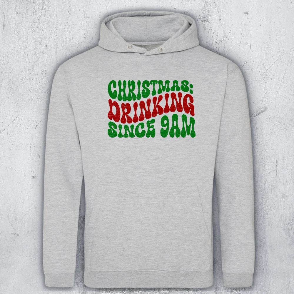 A light heather gray pullover hoodie with a graphic text print on the chest reading 'CHRISTMAS: DRINKING SINCE 9AM' in green and red lettering, displayed against a textured gray background.
