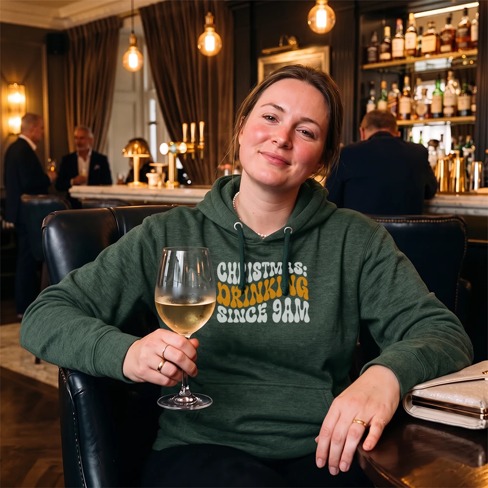 A woman sitting in a dimly lit bar or lounge holding a glass of white wine, wearing a dark green hoodie with the text 'CHRISTMAS IS: DRINKING SINCE 9AM' in yellow and white lettering.