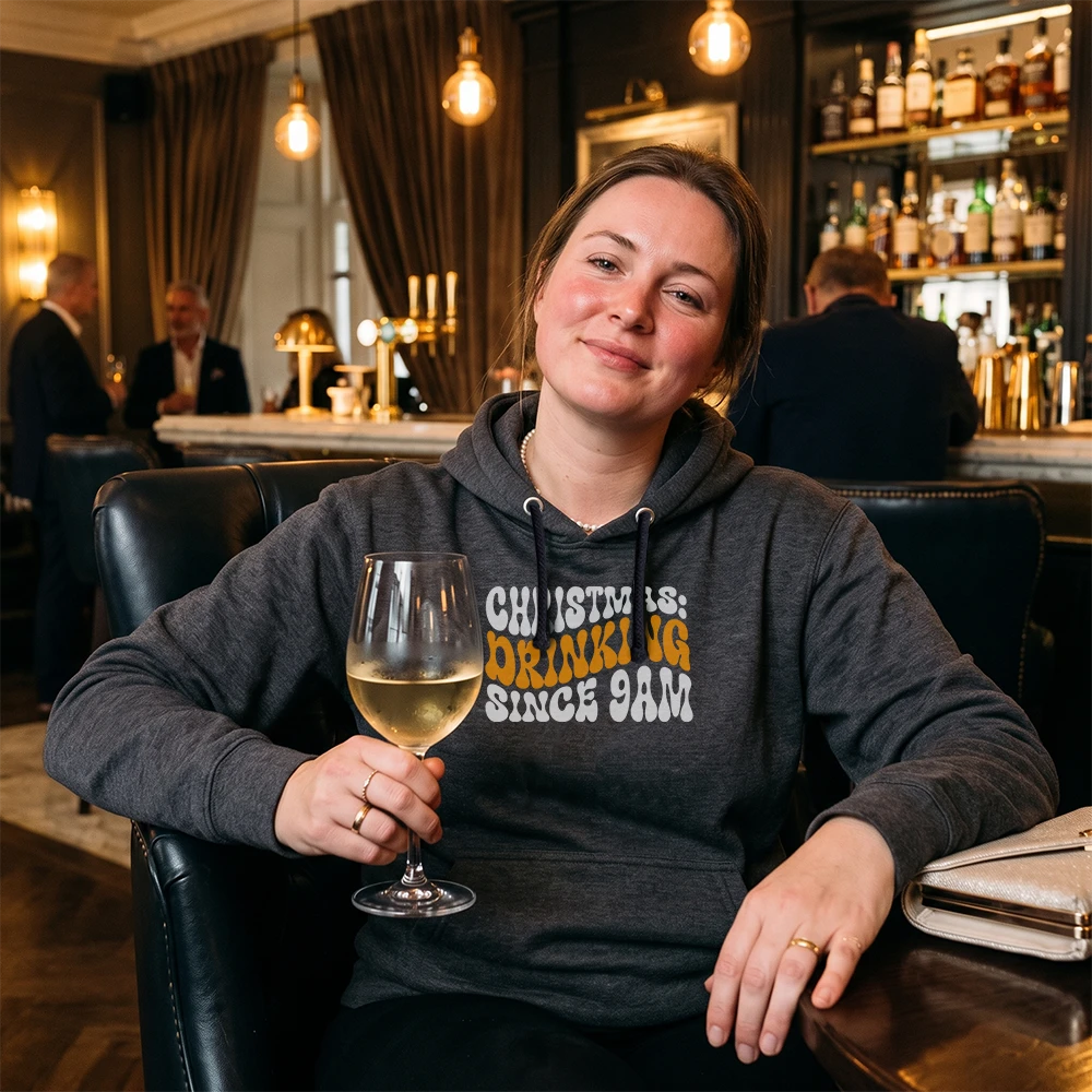 A woman sitting in a bar chair at a bar, holding a glass of white wine. She is wearing a dark gray hoodie with the text 'CHRISTMAS IS DRINKING SINCE 9AM' in white and orange lettering.