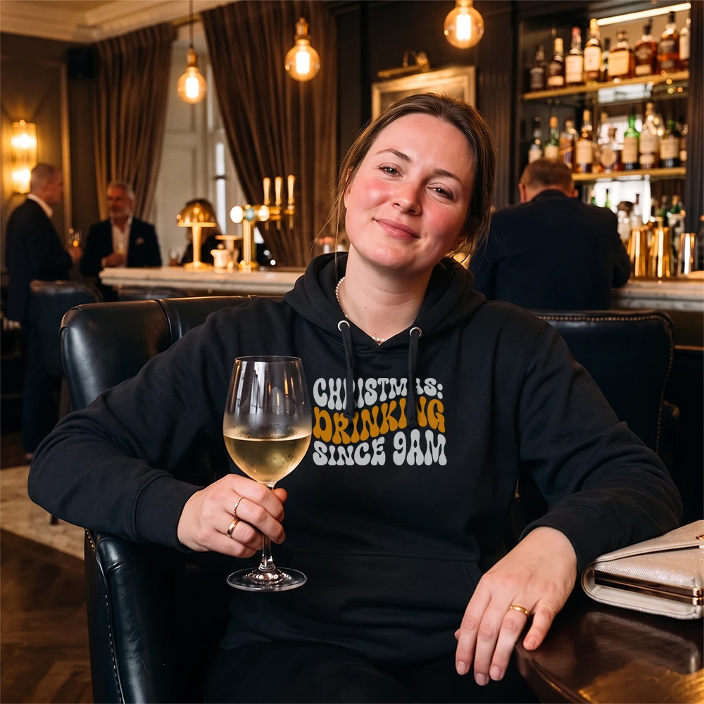 A woman sitting in a dimly lit bar or lounge, wearing a black hoodie with the text 'CHRIST IS: DRINKING SINCE 9AM' in white and orange lettering. She is holding a glass of white wine.