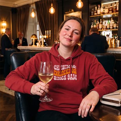 A woman in a red hoodie sits in a dimly lit bar holding a glass of white wine. The hoodie features the text 'CHRISTMAS IS: DRINKING SINCE 9AM' in white and orange lettering.