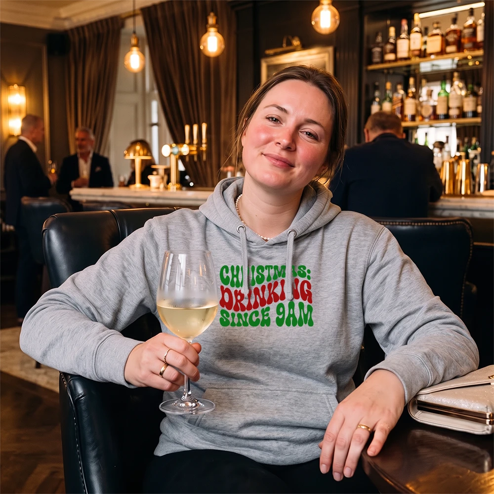A woman sitting in a bar or lounge setting wearing a light gray hoodie with the text 'CHRISTMAS IS: DRINKING SINCE 9AM' in green and red lettering. She is holding a glass of white wine.