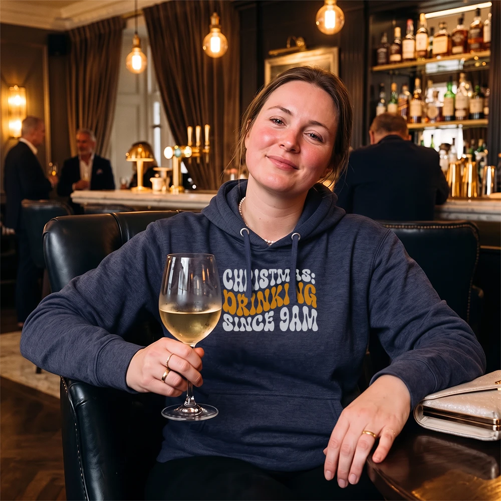 A woman sitting in a bar or lounge setting wearing a navy blue hoodie with the text 'CHRIST IS DRINKING SINCE 9AM' in white and orange. She is holding a glass of white wine.