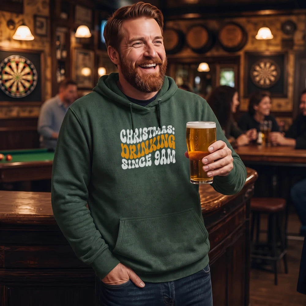 A smiling man in a green hoodie stands in a bar holding a pint of beer. The hoodie has a graphic print that reads 'CHRISTMAS DRINKING SINCE 9AM' in white and yellow text.