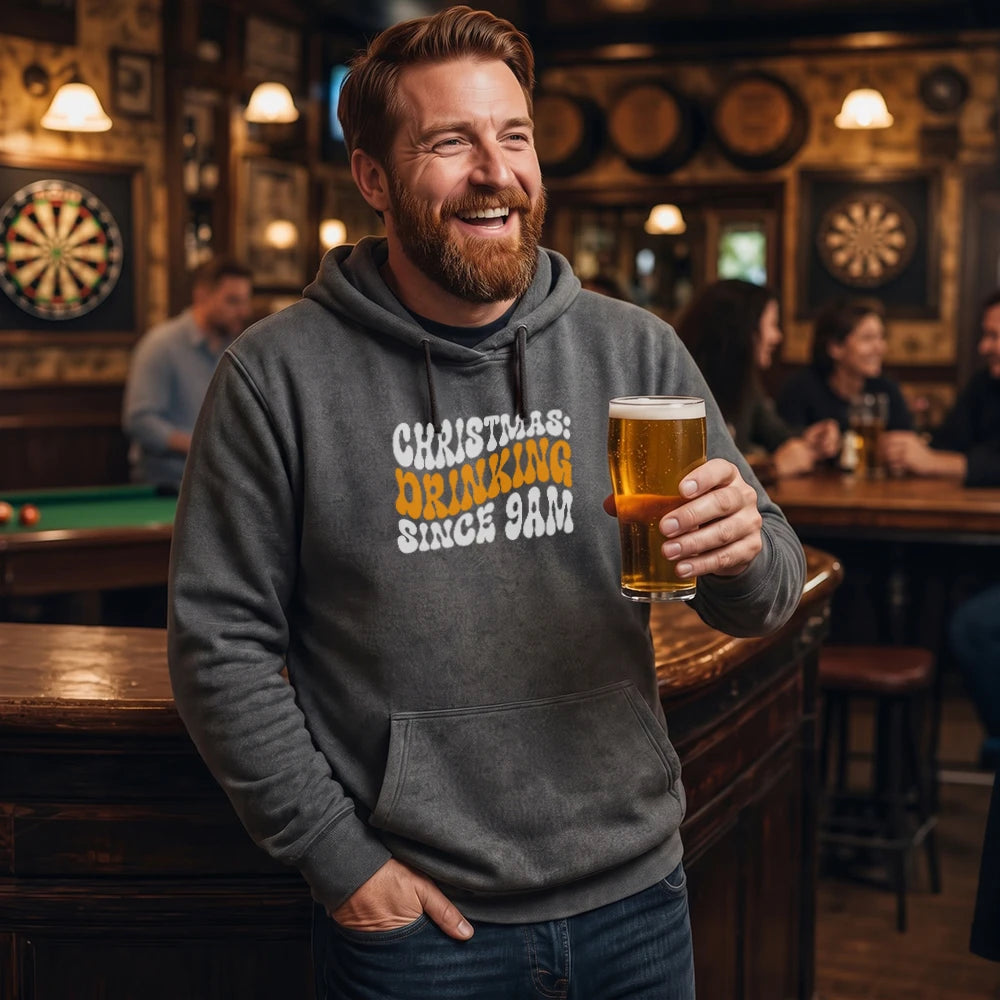 A smiling man in a bar wearing a dark gray hoodie with the text 'CHRISTMAS DRINKING SINCE 9AM' in white and orange, holding a glass of beer.