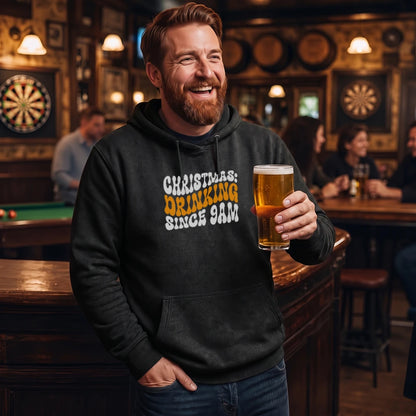 A smiling man in a bar wearing a black hoodie with the text 'CHRISTMAS: DRINKING SINCE 9AM' in white and yellow, holding a pint of beer.
