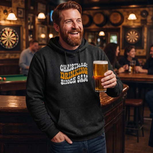 A smiling man in a bar wearing a black hoodie with the text 'CHRISTMAS: DRINKING SINCE 9AM' in white and yellow, holding a pint of beer.