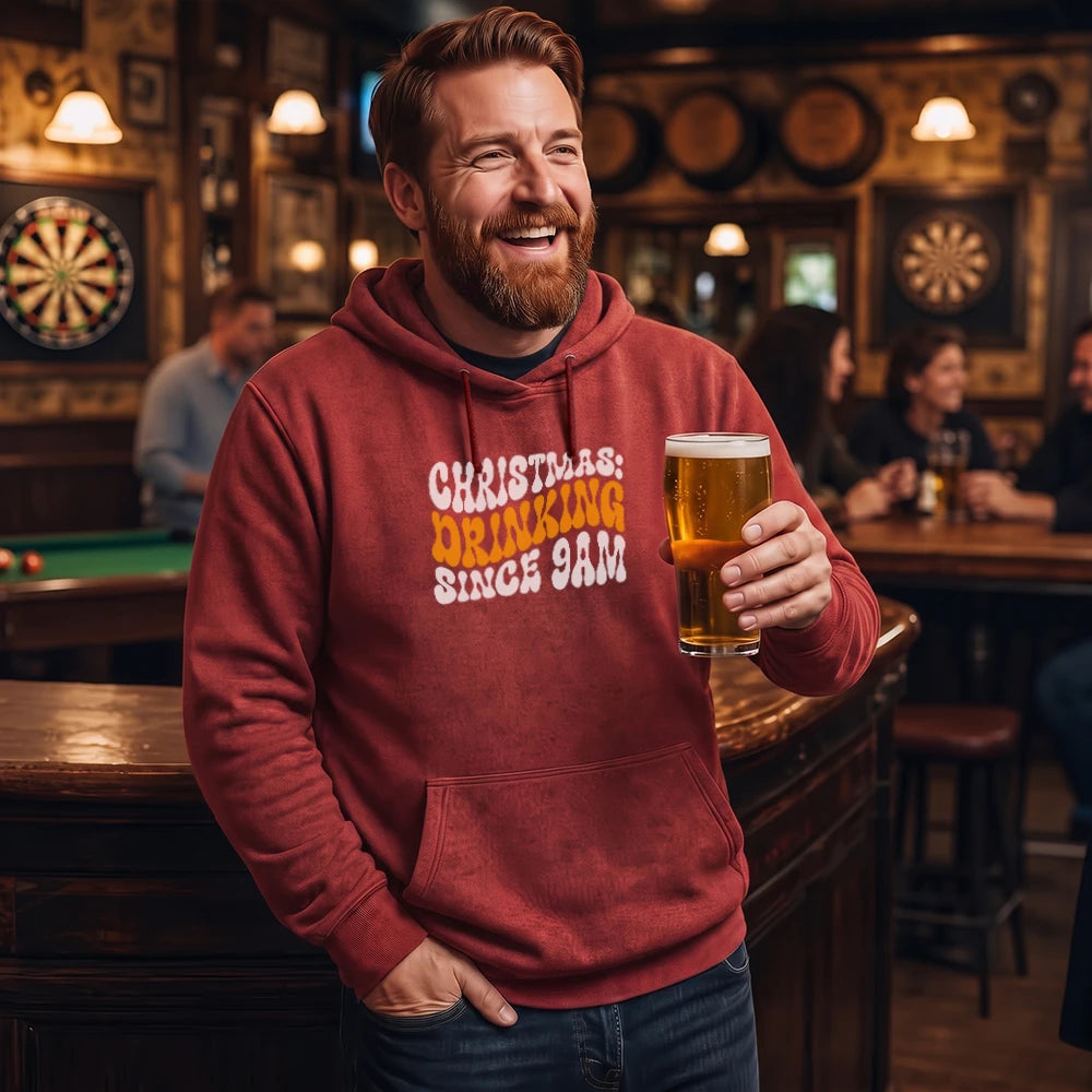 A smiling man in a bar wearing a red hoodie with the text 'CHRISTMAS DRINKING SINCE 9AM' in white and yellow, holding a pint of beer.