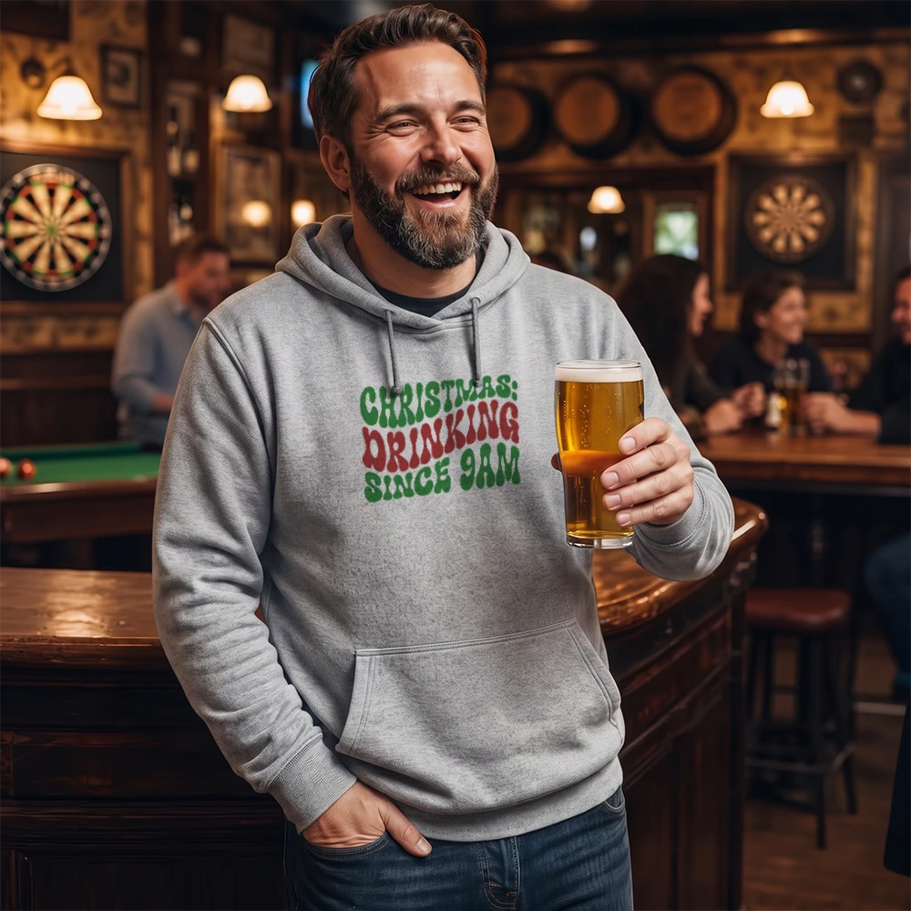 A man in a bar wearing a gray hoodie with a green and red Christmas-themed graphic that reads 'CHRISTMAS DRINKING SINCE 9AM', holding a pint of beer.