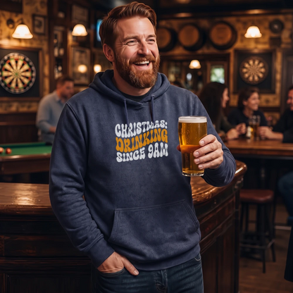 A smiling man in a bar holding a pint of beer, wearing a navy blue hoodie with the text 'CHRISTMAS: DRINKING SINCE 9AM' in white and yellow.