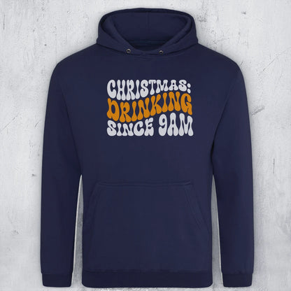 A navy blue pullover hoodie with a front graphic reading 'CHRISTMAS: DRINKING SINCE 9AM' in white and orange retro lettering, displayed against a light gray textured wall.