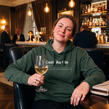A woman sitting in a dark leather chair at a bar, wearing a dark green hoodie with white text that reads 'Sauvignon Blank' and holding a glass of white wine.