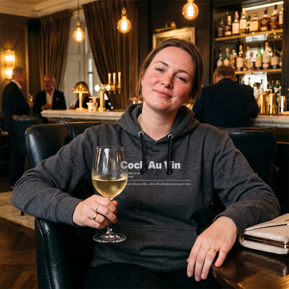 A woman sitting in a bar or lounge setting wearing a dark gray hoodie with white text that reads 'Sauvignon Blank' and holding a glass of white wine.