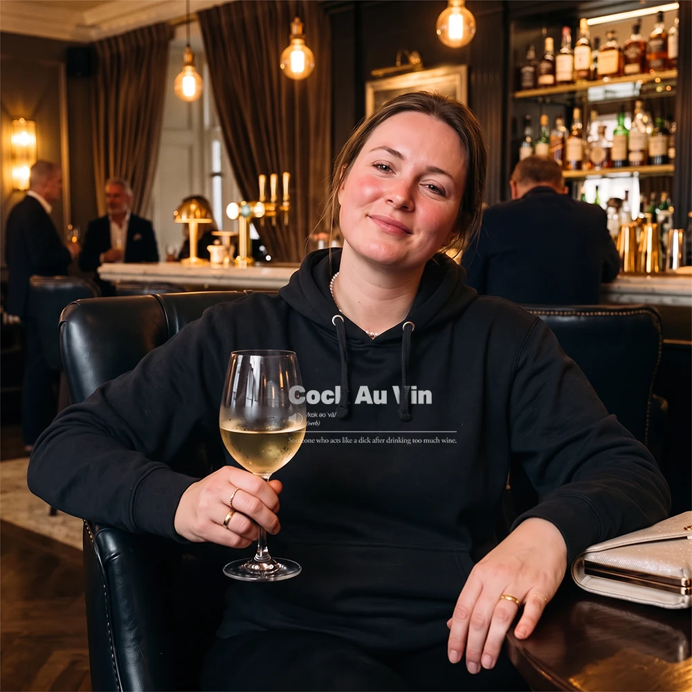 A woman sitting in a dimly lit bar or lounge, wearing a black hoodie with the text 'Sauvignon Blank' printed on the chest, and holding a glass of white wine.