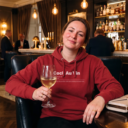 A woman wearing a red hoodie sits in a dimly lit bar holding a glass of white wine. The hoodie features white text that reads 'Cock Au Vin' 