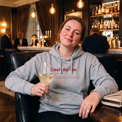 A woman sitting in a bar setting wearing a light gray hoodie with the text 'Sauvignon Blank' printed on the chest, holding a glass of white wine.