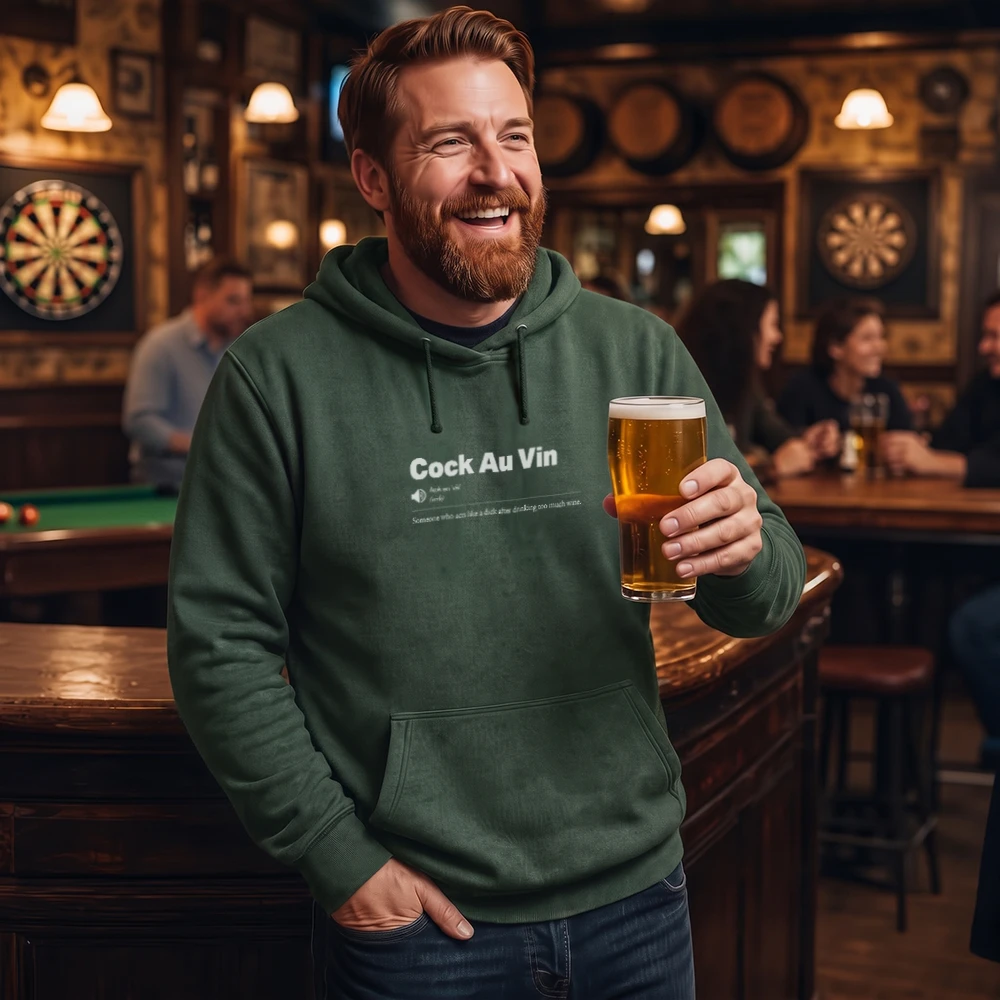 A smiling man in a bar wearing a dark green hoodie with the text 'Cock Au Vin' printed on the chest, holding a pint of beer.