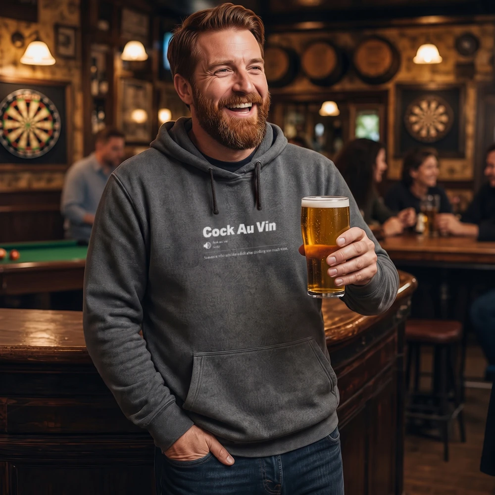 A smiling man in a bar wearing a dark gray hoodie with the text 'Cock Au Vin' and holding a pint of beer.