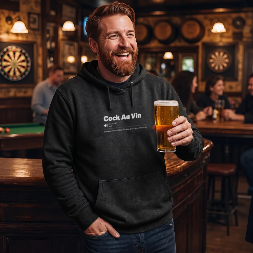 A smiling man in a bar wearing a black hoodie with white text that reads 'Cock Au Vin' and holding a pint of beer.